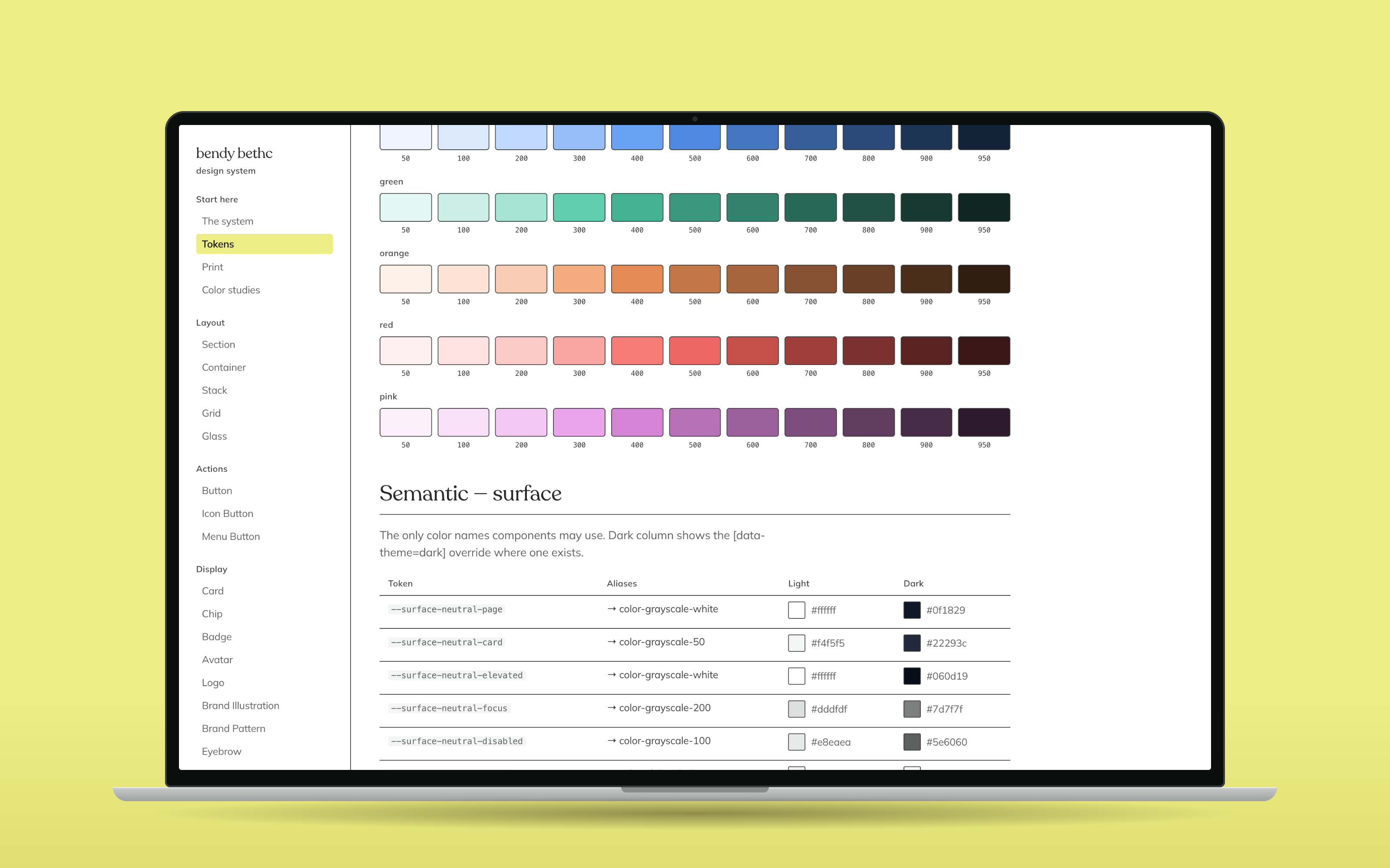 A laptop on a pale yellow ground showing the Tokens page of the design system showcase: colour primitive swatches and the semantic surface table with light and dark values side by side.